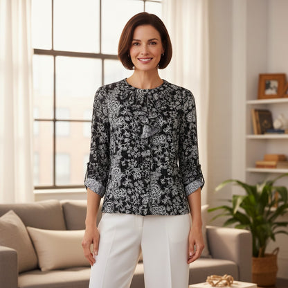 A woman stands in a bright living room wearing a black and white floral patterned blouse with three-quarter sleeves and white pants.