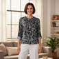 A woman stands in a bright living room wearing a black and white floral patterned blouse with three-quarter sleeves and white pants.