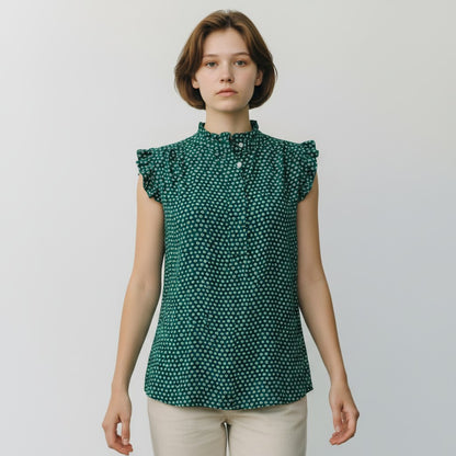 A woman modeling a sleeveless green blouse with a dense small white polka dot pattern, ruffled cap sleeves, and a high ruffled collar, paired with light beige trousers, against a plain light gray background.