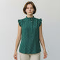 A woman modeling a sleeveless green blouse with a dense small white polka dot pattern, ruffled cap sleeves, and a high ruffled collar, paired with light beige trousers, against a plain light gray background.