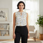 A woman stands in a bright, modern living room wearing a sleeveless white blouse with a delicate multicolor floral print and a keyhole tie neckline, paired with black trousers.