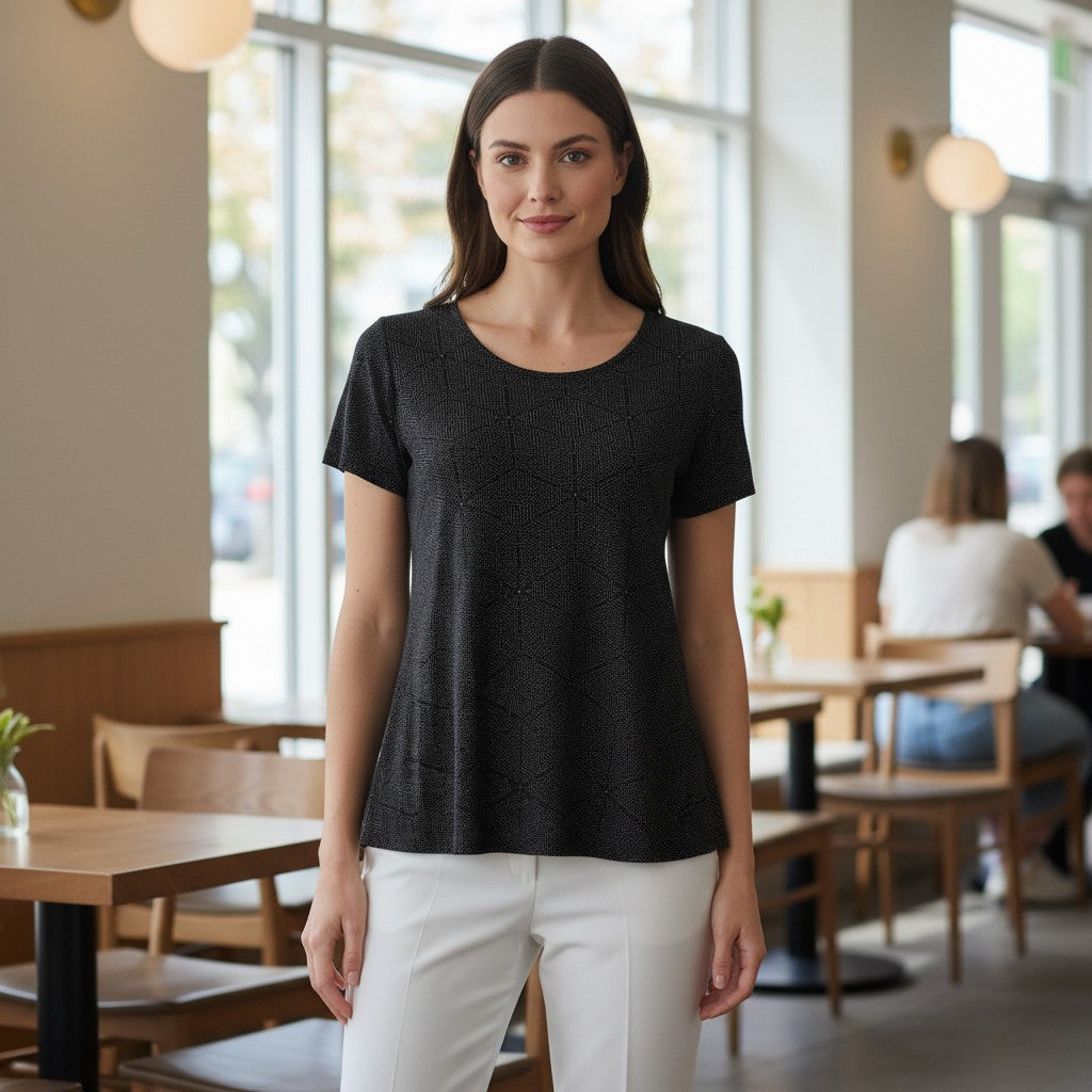 A woman stands in a bright cafe wearing a black short-sleeve textured knit top and white pants.
