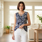 A woman modeling a sleeveless navy blue blouse with a white floral and paisley pattern, featuring a lace-up V-neckline, paired with white pants in a bright, modern living room.