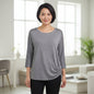 A woman wearing a heather gray three-quarter sleeve top with lace shoulder panels and a side twist detail, paired with black pants, standing in a bright, modern living room.
