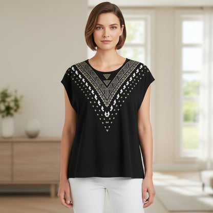 A woman modeling a black short-sleeve top with a geometric embroidered yoke in white and metallic gold, paired with white pants, set against a bright, softly blurred indoor background.