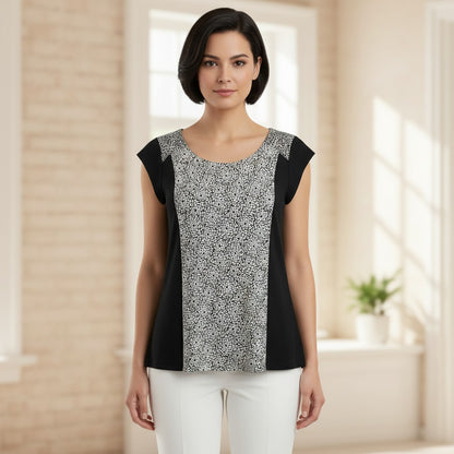 A woman modeling a short-sleeve scoop-neck top with a black and white abstract patterned center panel and solid black side panels, paired with white pants in a bright indoor setting.