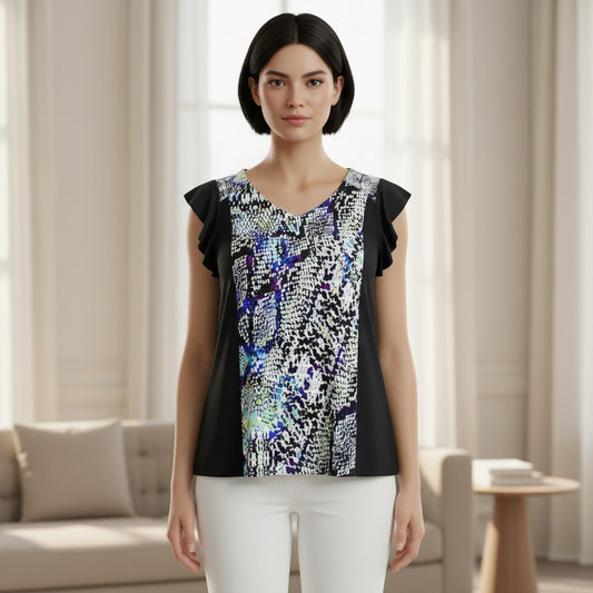 A woman stands in a bright living room wearing a sleeveless top with black side panels and a colorful abstract print on the front, paired with white pants.