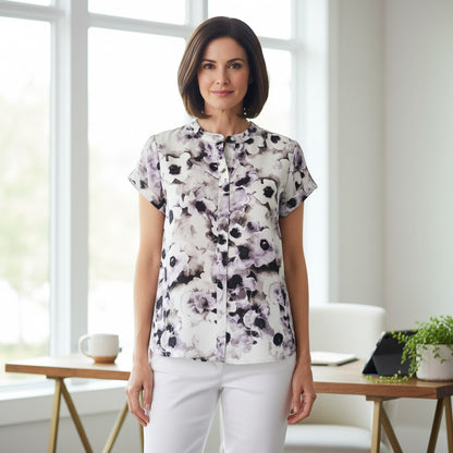 A woman stands indoors wearing a short-sleeve button-front blouse with a white base and a black and gray floral print, paired with white pants. The setting is a bright room with large windows, a wooden table, and a chair in the background.