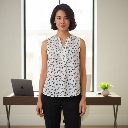 A woman stands indoors wearing a sleeveless white blouse with a black abstract print and black pants. The setting includes a desk with a laptop and a small plant in front of a bright window.