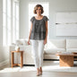 A woman stands in a bright, modern living room wearing a black and white patterned tiered short-sleeve blouse and white pants.