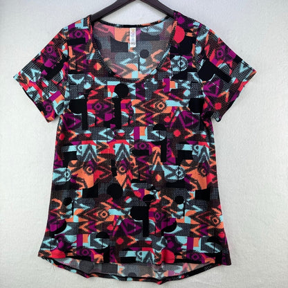 A women's short-sleeve V-neck top with a vibrant geometric and tribal-inspired print in black, pink, orange, and light blue, hanging on a dark hanger against a textured off-white wall.
