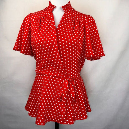 A red short-sleeve blouse with white polka dots, featuring a high stand collar, ruffled sleeves, and a matching fabric tie belt at the waist, displayed on a mannequin against a light gray fabric backdrop.