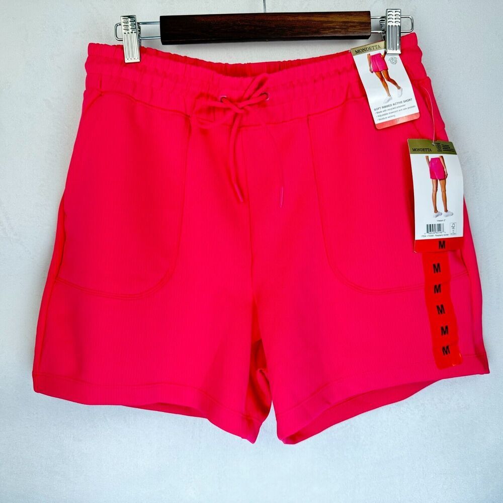 Bright pink athletic shorts with an elastic drawstring waist and side pockets, hanging on a wooden hanger with retail tags attached against a light gray wall.