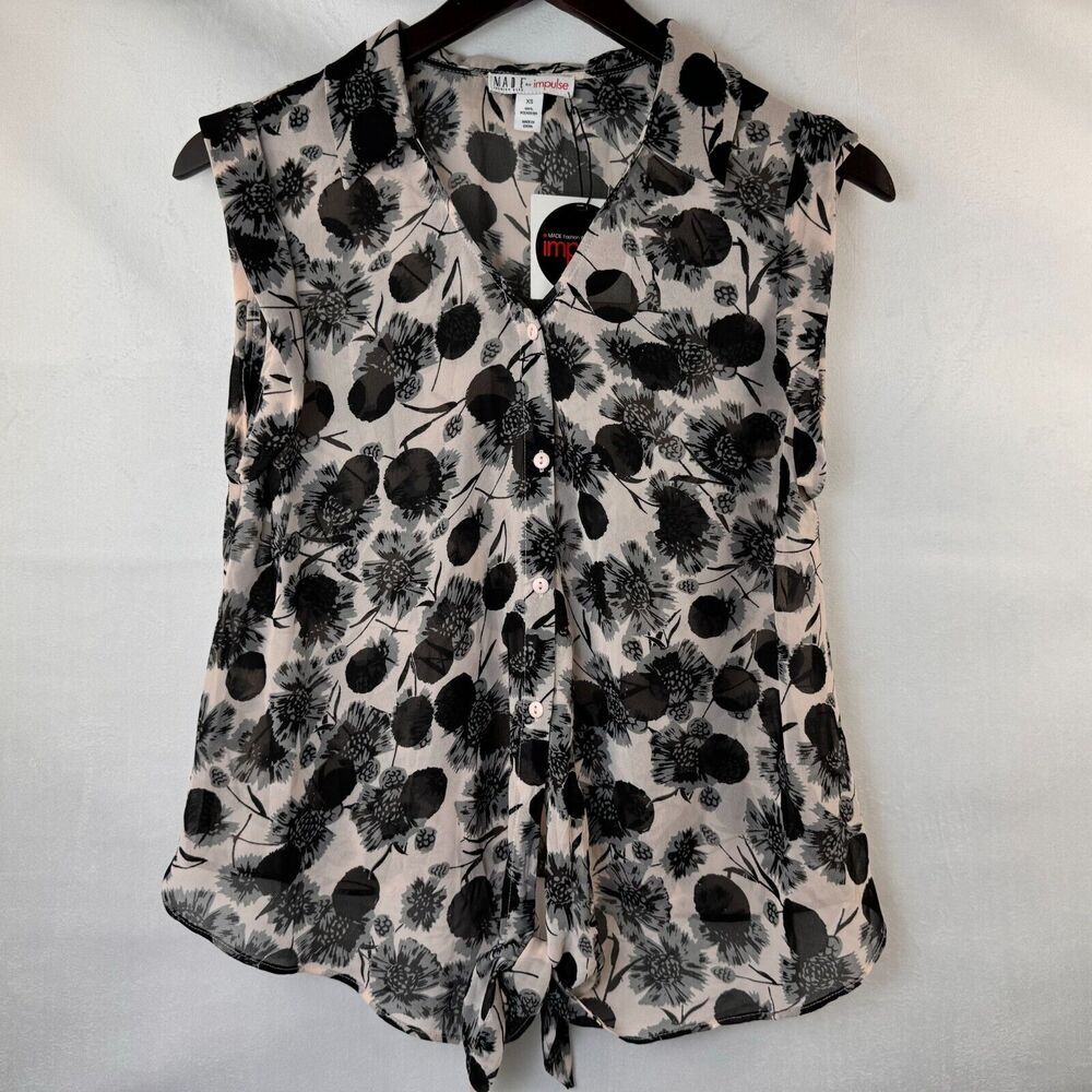 A sleeveless button-front blouse with a black floral and botanical print on a light beige background, hanging on a black hanger against a white fabric backdrop.