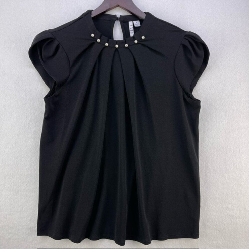A black short-sleeve blouse with a pleated front and decorative pearl-like buttons at the neckline, hanging on a wooden hanger against a light gray textured wall.