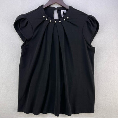A black short-sleeve blouse with a pleated front and decorative pearl-like buttons at the neckline, hanging on a wooden hanger against a light gray textured wall.