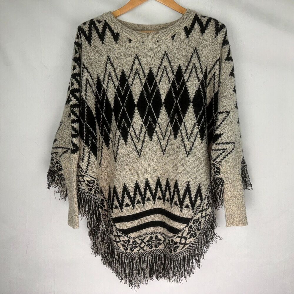 A knit poncho sweater with a black and beige geometric diamond and zigzag pattern, featuring fringe trim along the hem and sleeves, hanging on a wooden hanger against a white fabric background.
