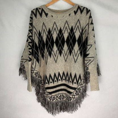 A knit poncho sweater with a black and beige geometric diamond and zigzag pattern, featuring fringe trim along the hem and sleeves, hanging on a wooden hanger against a white fabric background.