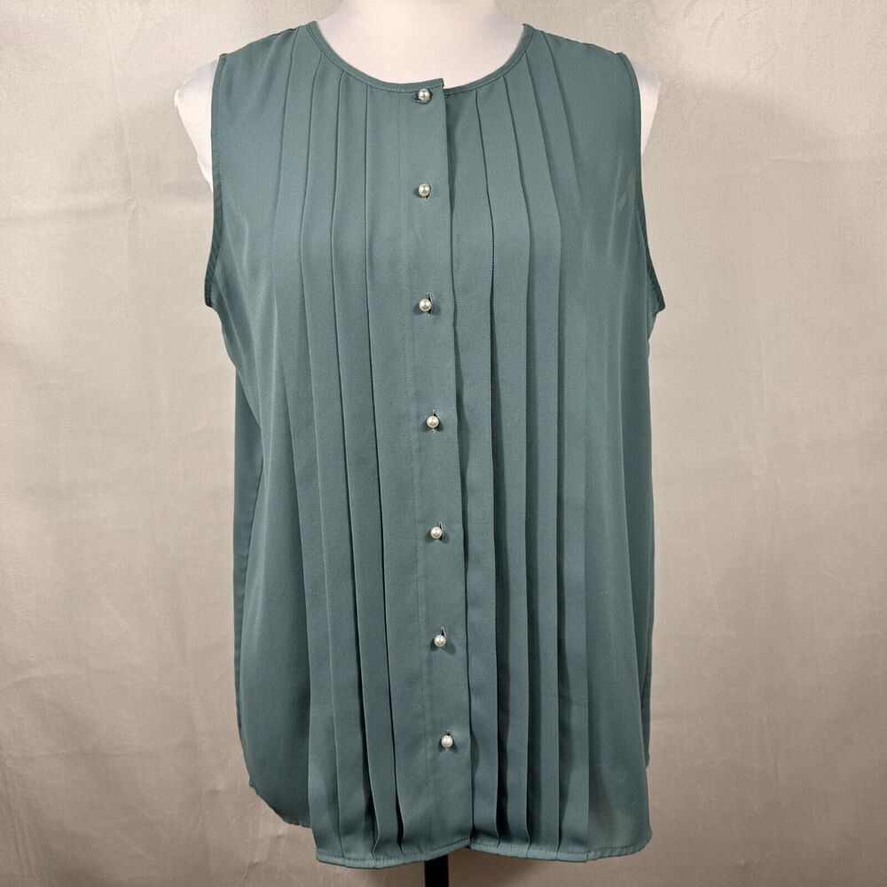 A sleeveless, scoop-neck blouse in a muted teal-green color, featuring vertical pleats and a front button closure with pearl-like buttons, displayed on a white mannequin torso against a neutral beige fabric backdrop.