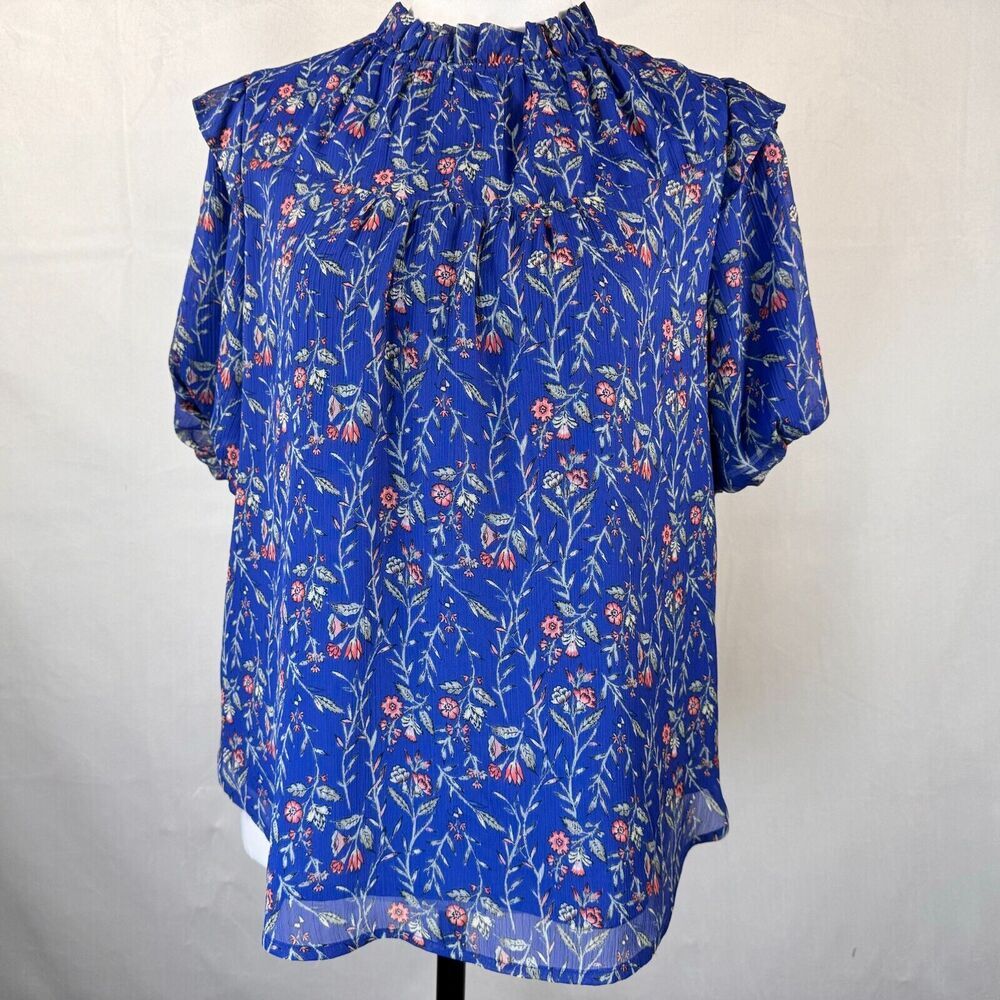 A blue short-sleeve blouse with a dense floral and leaf print in pink, red, and white, displayed on a mannequin torso against a light gray fabric backdrop.