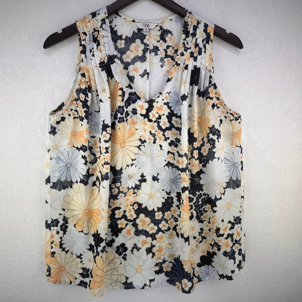 A sleeveless V-neck blouse with a floral print, hanging on a dark wooden hanger against a light textured wall. The print features large white and yellow flowers with dark navy/black and gray accents.