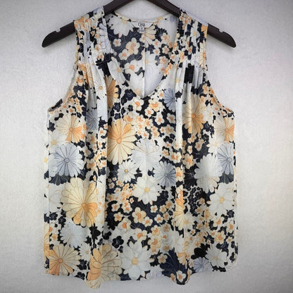 A sleeveless V-neck blouse with a floral print, hanging on a dark wooden hanger against a light textured wall. The print features large white and yellow flowers with dark navy/black and gray accents.