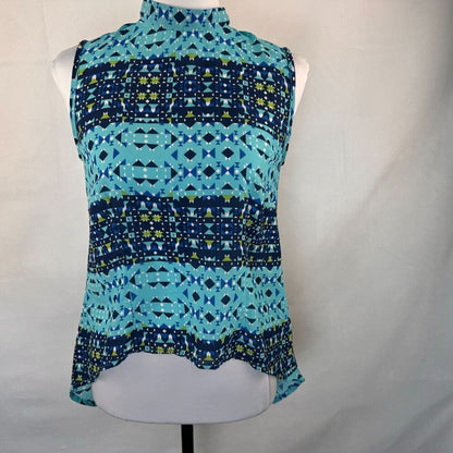 A sleeveless mock-neck blouse with a geometric pattern in shades of blue, teal, and lime green, displayed on a white mannequin torso against a white fabric backdrop.