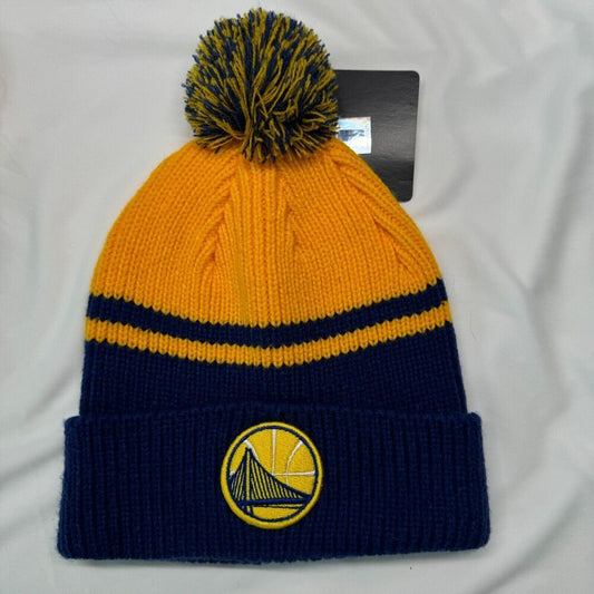A Golden State Warriors knit beanie hat in yellow and navy blue, featuring a matching yellow and navy pom-pom and the team logo embroidered on the front cuff. A black retail tag is attached near the top, and the hat is laid flat on a white fabric background.