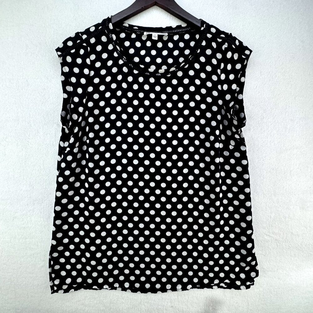 A sleeveless black top with a white polka dot pattern, hanging on a dark wooden hanger against a textured white wall.