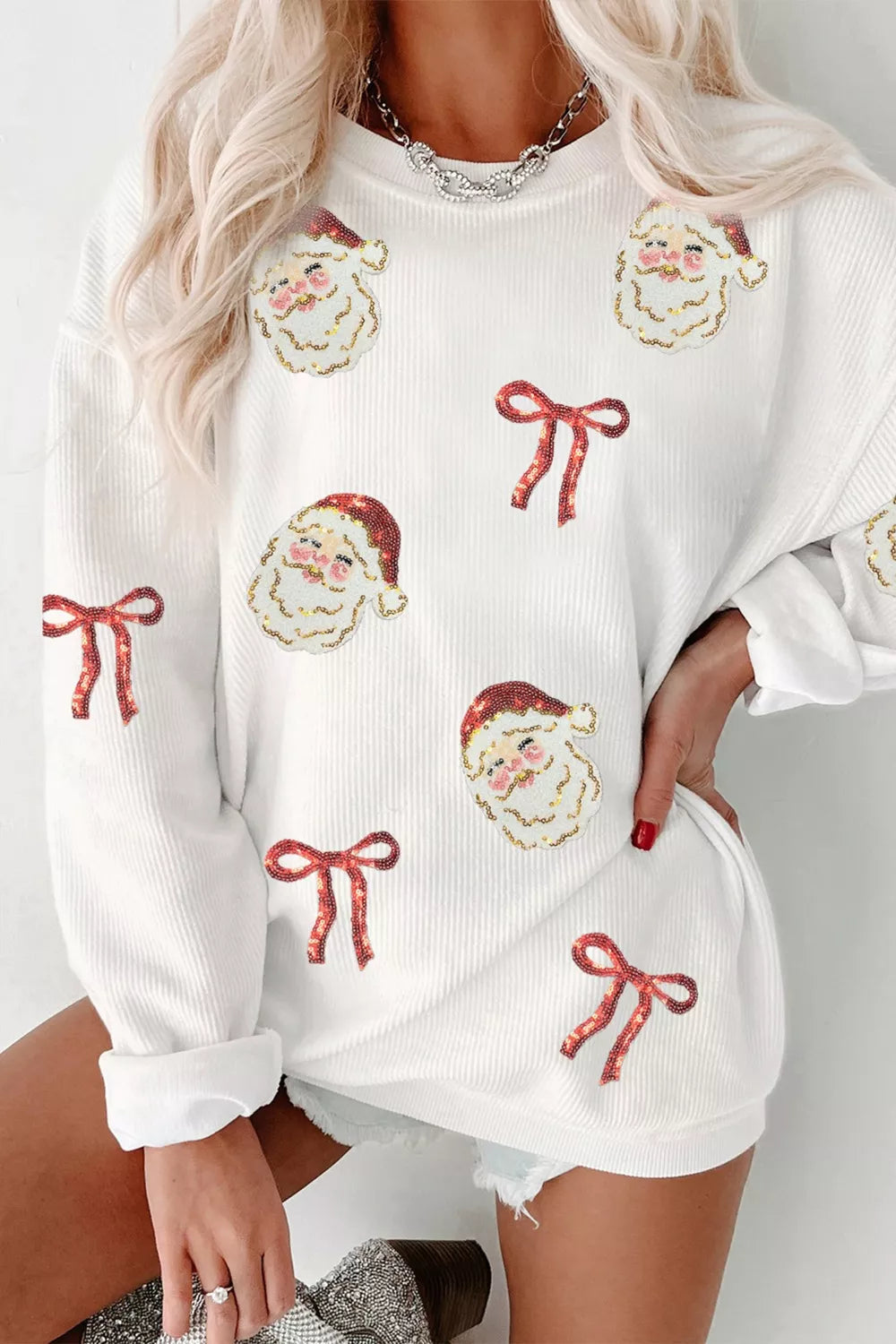 Woman wearing a white Simply Love Christmas sweatshirt decorated with sequin Santa faces and red ribbon bows, featuring long sleeves and a relaxed fit.