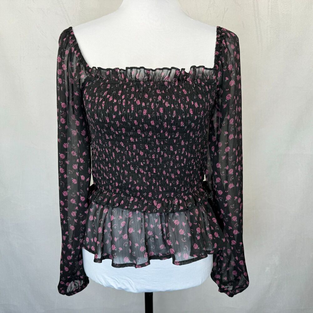 A black sheer long-sleeve blouse with a pink floral pattern, displayed on a white mannequin torso. The top features a smocked bodice, square neckline, and a ruffled peplum hem.