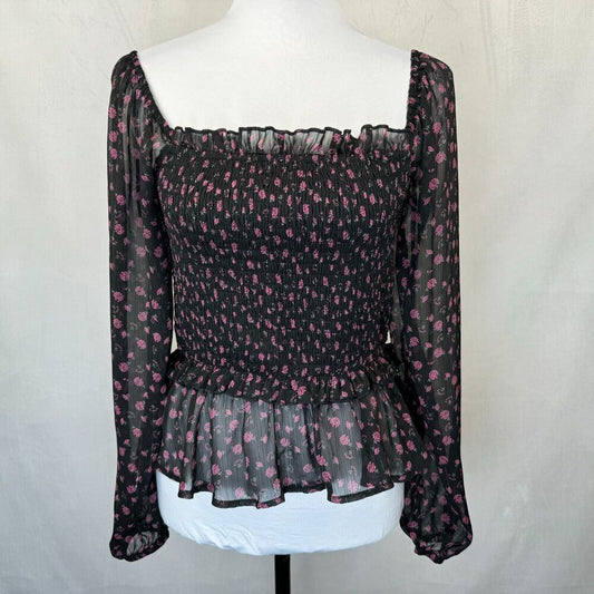A black sheer long-sleeve blouse with a pink floral pattern, displayed on a white mannequin torso. The top features a smocked bodice, square neckline, and a ruffled peplum hem.