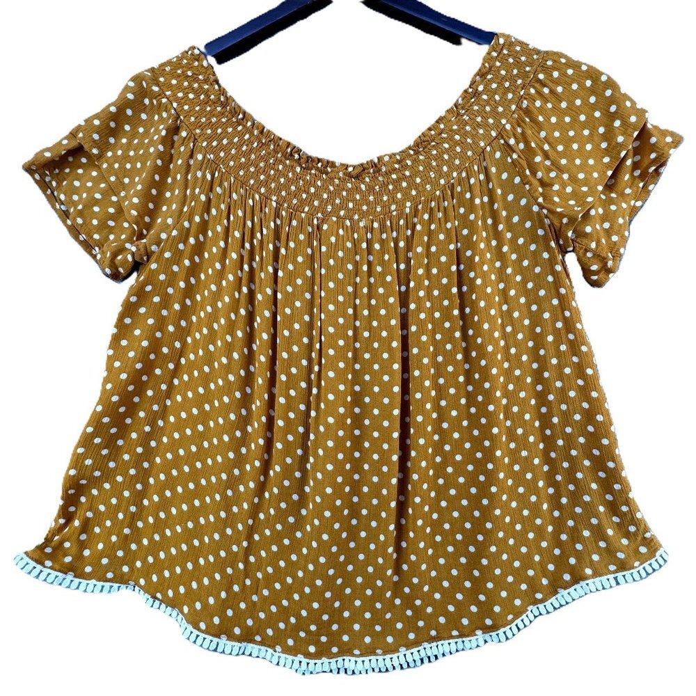 A women's short-sleeve top in mustard yellow with a white polka dot pattern, featuring an off-the-shoulder neckline and a scalloped ruffle trim along the hem, hanging on a black hanger against a white background.