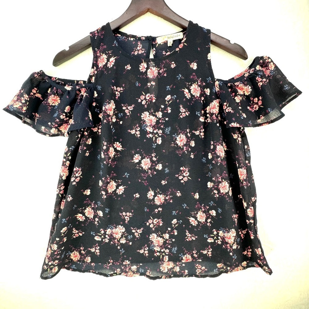 A sleeveless cold-shoulder blouse with a ruffled sleeve and an all-over floral print, hanging on a wooden hanger against a plain light background.