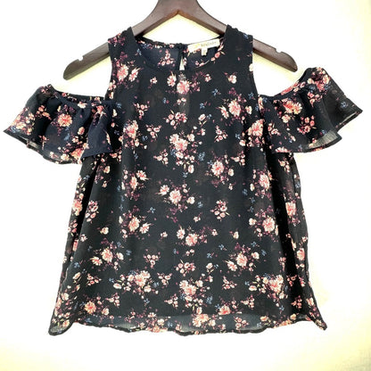 A sleeveless cold-shoulder blouse with a ruffled sleeve and an all-over floral print, hanging on a wooden hanger against a plain light background.