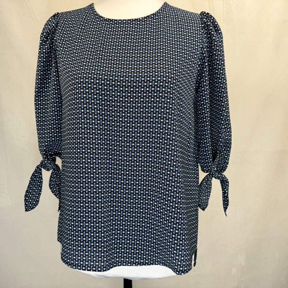 A women's long-sleeve blouse with a dense small-scale white geometric pattern on a dark navy or black base, featuring puffed sleeves with side tie details, displayed on a mannequin torso against a neutral beige backdrop.