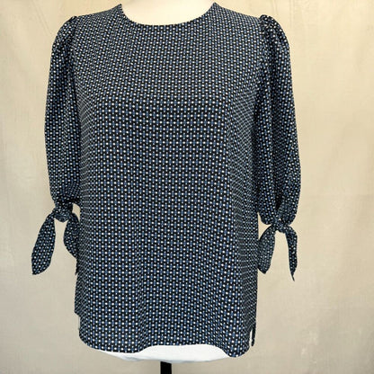 A women's long-sleeve blouse with a dense small-scale white geometric pattern on a dark navy or black base, featuring puffed sleeves with side tie details, displayed on a mannequin torso against a neutral beige backdrop.