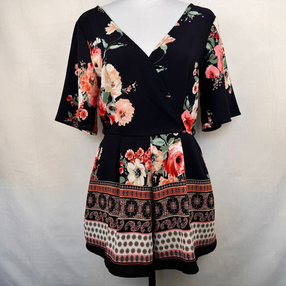A women's short-sleeve wrap dress displayed on a mannequin against a light gray fabric backdrop. The dress has a black base with a vibrant floral print in pink, red, and cream, and features a decorative border pattern on the skirt.