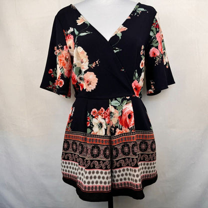 A women's short-sleeve wrap dress displayed on a mannequin against a light gray fabric backdrop. The dress has a black base with a vibrant floral print in pink, red, and cream, and features a decorative border pattern on the skirt.