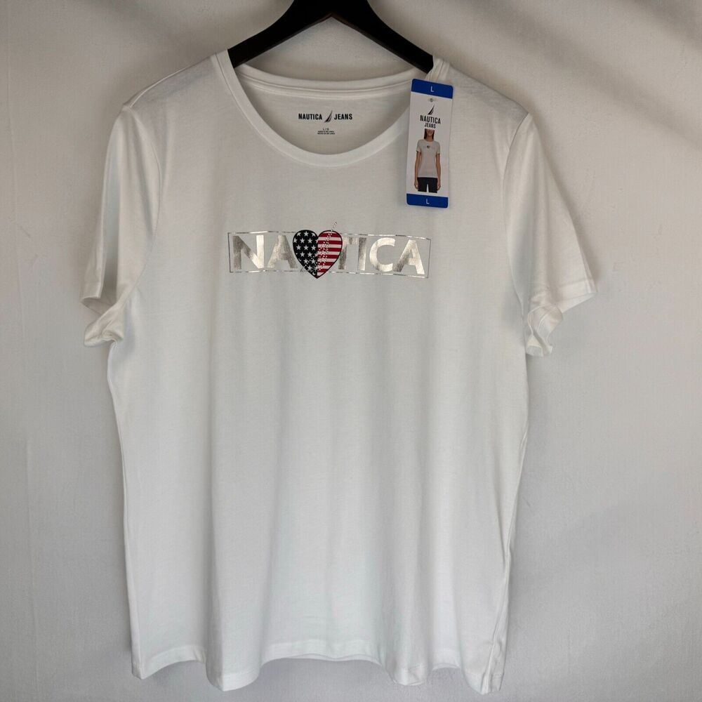 A white short-sleeve crewneck t-shirt by Nautica, hanging on a black hanger against a light gray wall. The front features a graphic spelling 'NAUTICA' with a heart made of an American flag pattern (red, white, and blue).