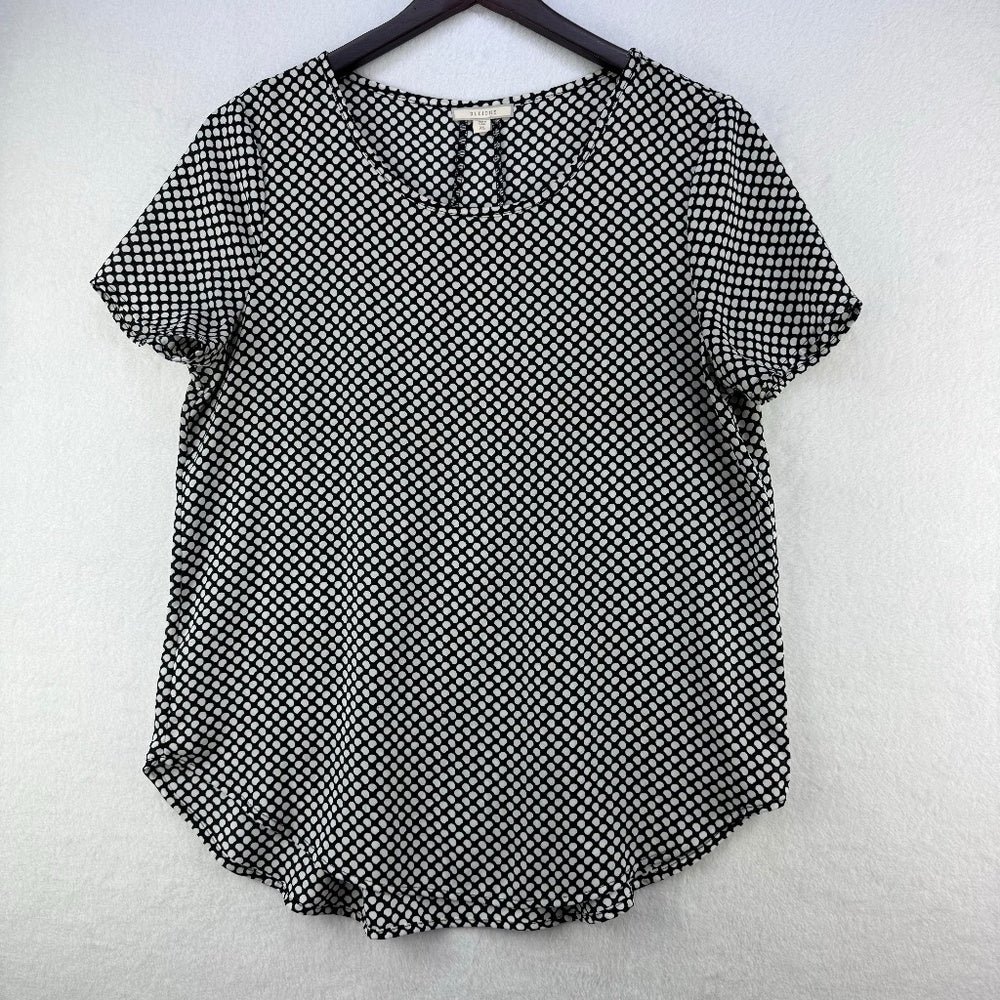 A short-sleeve black and white patterned top hanging on a black hanger against a light gray textured wall.