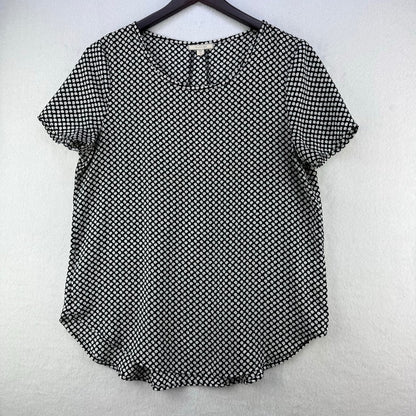 A short-sleeve black and white patterned top hanging on a black hanger against a light gray textured wall.