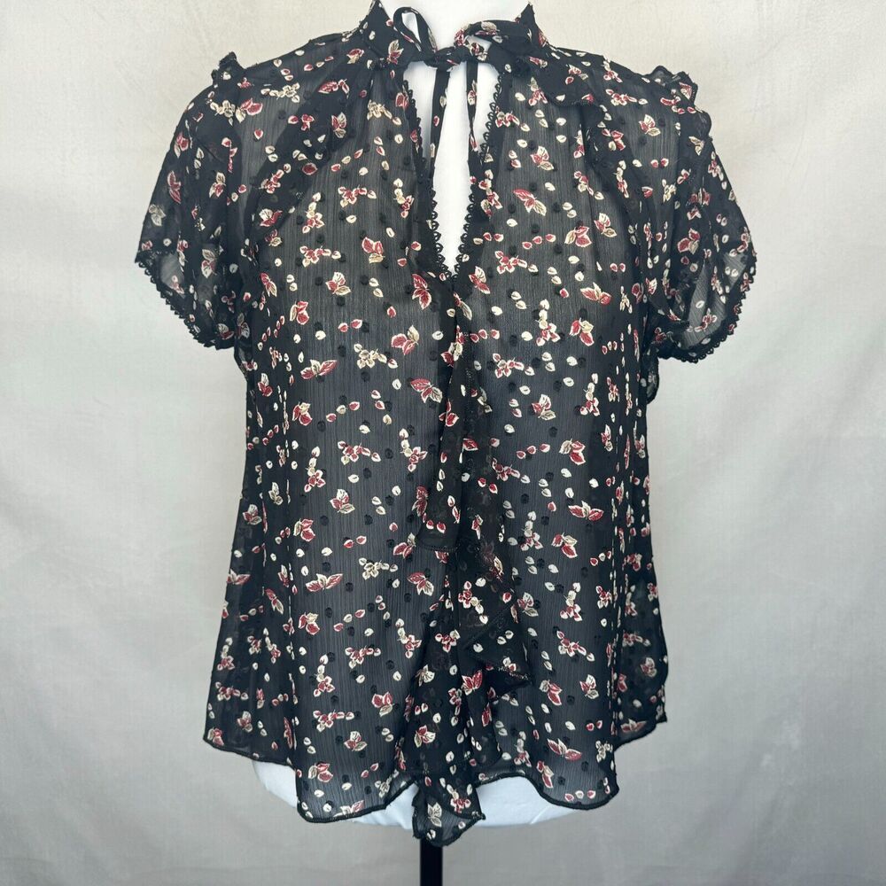 A sheer black short-sleeve blouse with a red and white floral print, featuring a tie-neck detail and ruffled trim, displayed on a mannequin torso against a light gray fabric backdrop.