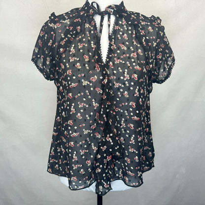 A sheer black short-sleeve blouse with a red and white floral print, featuring a tie-neck detail and ruffled trim, displayed on a mannequin torso against a light gray fabric backdrop.