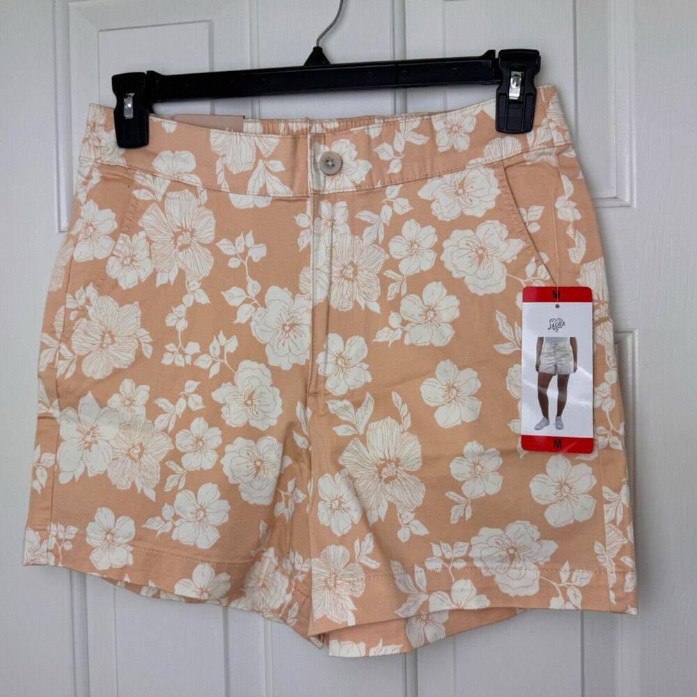 A pair of women's shorts with a peach and white floral print, hanging on a black hanger against a white paneled door. A red and white Gildan brand tag is attached to the right side.