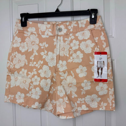 A pair of women's shorts with a peach and white floral print, hanging on a black hanger against a white paneled door. A red and white Gildan brand tag is attached to the right side.