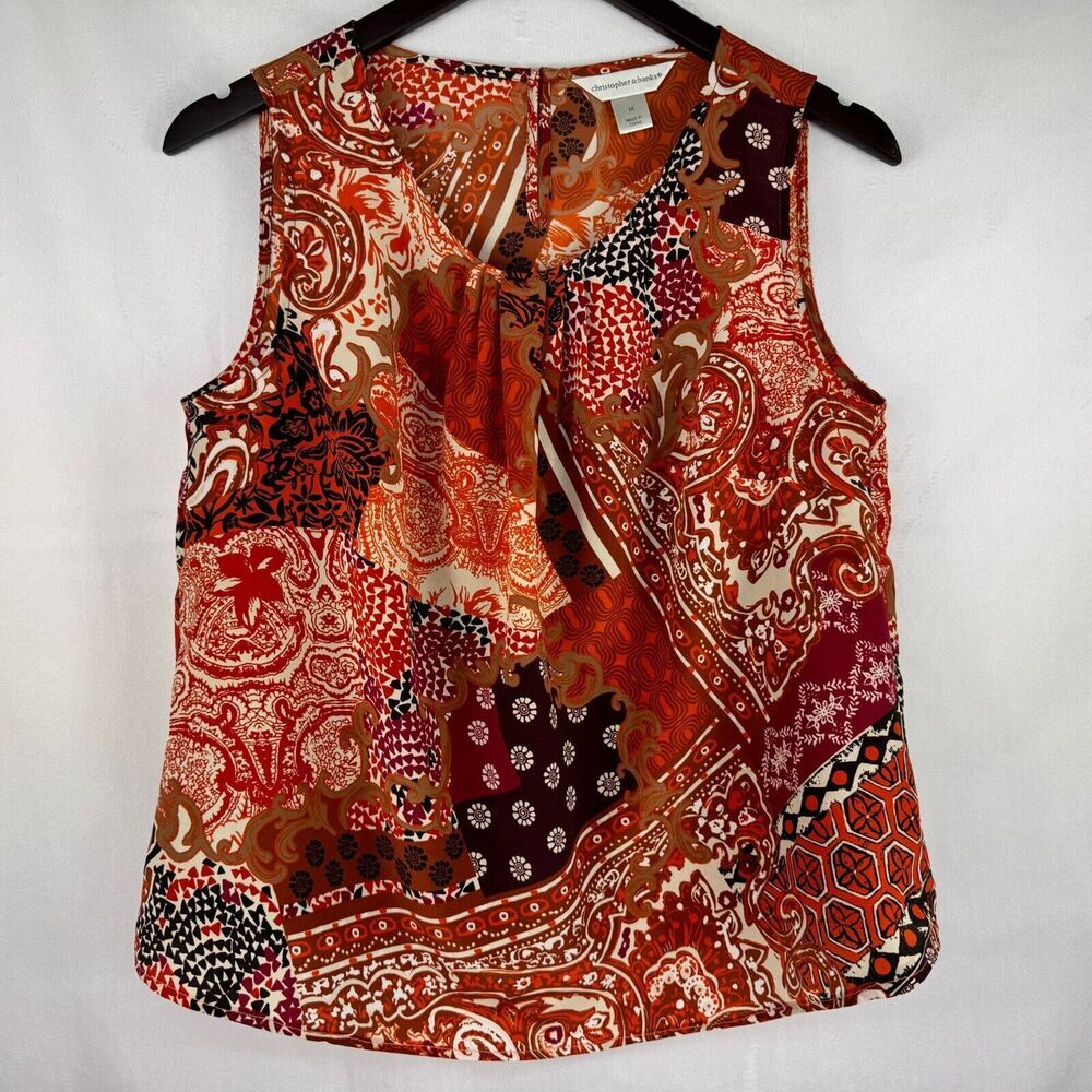 A sleeveless wrap-style blouse with a ruffled tie neckline, featuring a busy patchwork paisley and floral print in red, orange, brown, black, and cream, hanging on a black hanger against a white background.