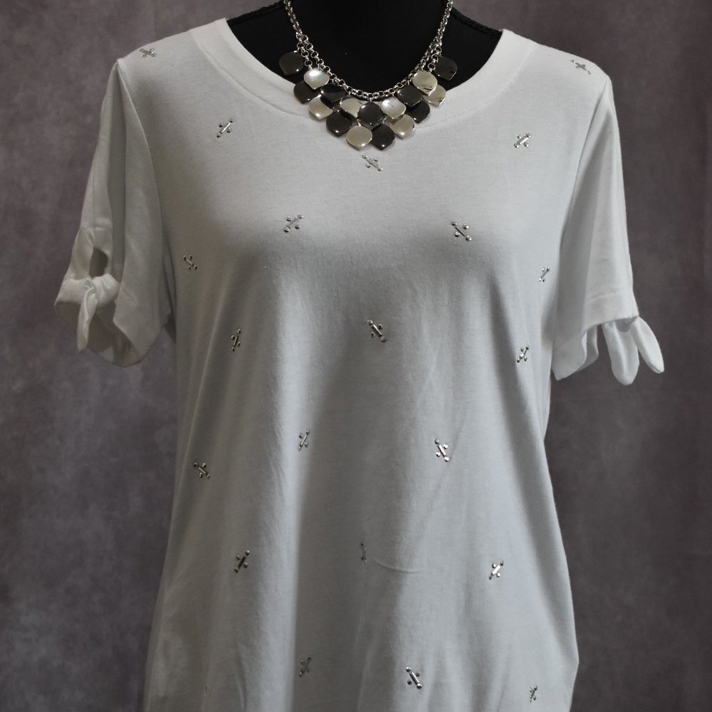 A white short-sleeve tunic top with small silver cross embellishments and ruffled tie details on the sleeves, displayed on a black mannequin torso with a silver statement necklace against a textured gray background.