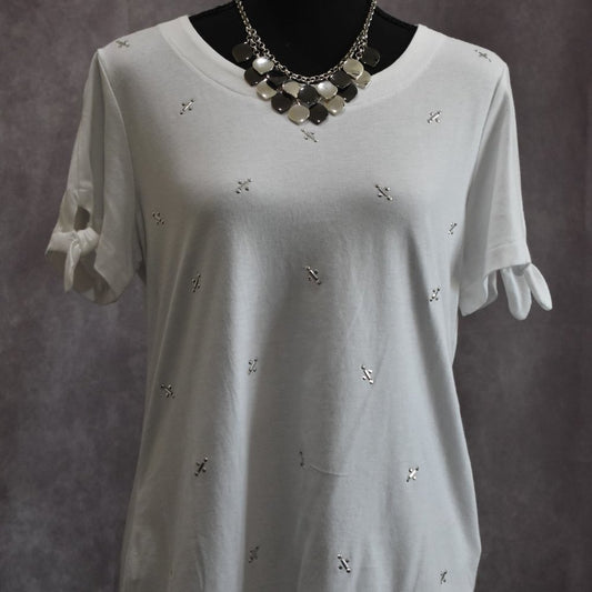 A white short-sleeve tunic top with small silver cross embellishments and ruffled tie details on the sleeves, displayed on a black mannequin torso with a silver statement necklace against a textured gray background.