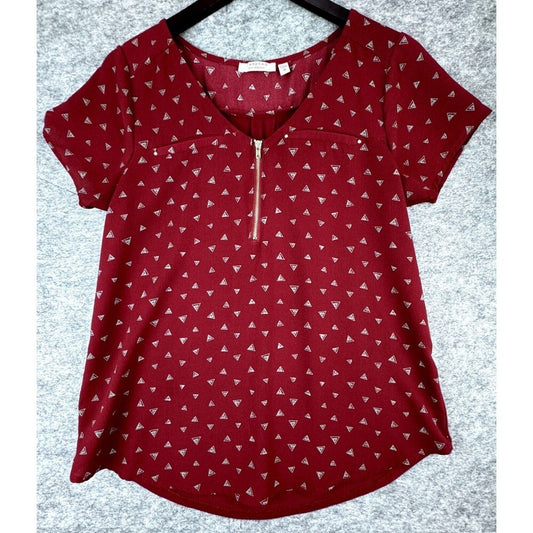 A red short-sleeve top with a small white geometric triangle pattern, featuring a split V-neckline with a gold-tone zipper and a chest pocket, shown on a black hanger against a gray textured background.